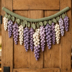 Peut inclure: Une guirlande de glycine au crochet faite à la main, dans des tons de violet et de crème, avec des feuilles vertes et une vigne verte. La guirlande est exposée contre une porte en bois, mettant en valeur les détails.