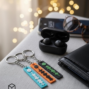Könnte beinhalten: Drei Schlüsselanhänger mit Spotify-Code-Designs in Blau, Orange und Schwarz, jeweils an einem silbernen Schlüsselring befestigt. Ein Paar schwarze kabellose Ohrhörer in einem Ladeetui, eine Brieftasche und eine Sonnenbrille sind ebenfalls zu sehen.