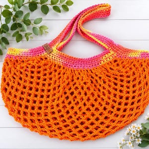 May include: A crochet market bag featuring a vibrant orange open-mesh body and a solid pink and yellow variegated top border. The bag includes two matching crochet handles and a small brown rectangular leather-like tag attached to the upper rim.