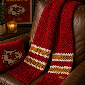 May include: A red scarf with white and gold stripes and fringed ends, draped over a brown leather armchair. The scarf has a textured weave. A red cushion with a team logo is also visible.