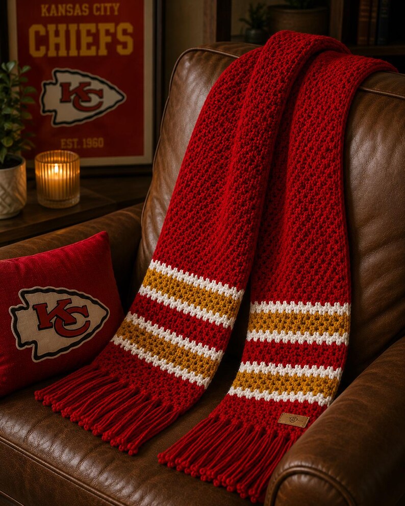 May include: A red scarf with white and gold stripes and fringed ends, draped over a brown leather armchair. The scarf has a textured weave. A red cushion with a team logo is also visible.