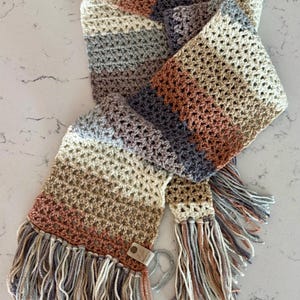 May include: A handmade crochet scarf in a neutral color palette of beige, gray, and muted rose. The scarf features a fringed edge and a tag. The open weave design adds texture and visual interest to this cozy accessory.