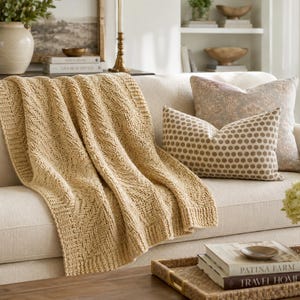 May include: A beige knit throw blanket with a textured chevron pattern draped over a light-colored sofa. The blanket features a thick ribbed border and a soft, woven appearance. Two decorative throw pillows are placed on the sofa next to the blanket.