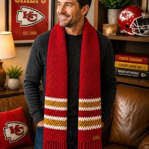 May include: A red and gold striped scarf with tassels. The scarf is worn by a person. The background includes a framed poster that says "Kansas City Chiefs" and a red cushion with the Chiefs logo.