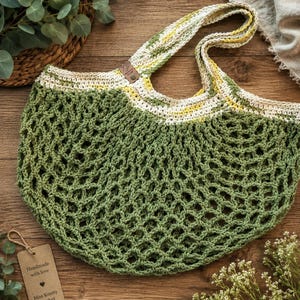 May include: A crochet market bag featuring a sage green net body and a variegated cream, yellow, and green handle. A small brown leather-like tag is attached to the strap. A rectangular cardboard tag reads Handmade with love, a heart, and Miss Knotty Crochet.
