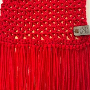 May include: A close-up of a vibrant red crocheted scarf with a fringe. The scarf features an open weave pattern. A small leather tag with the text "Miss Pretty Crochet" is attached to the scarf.