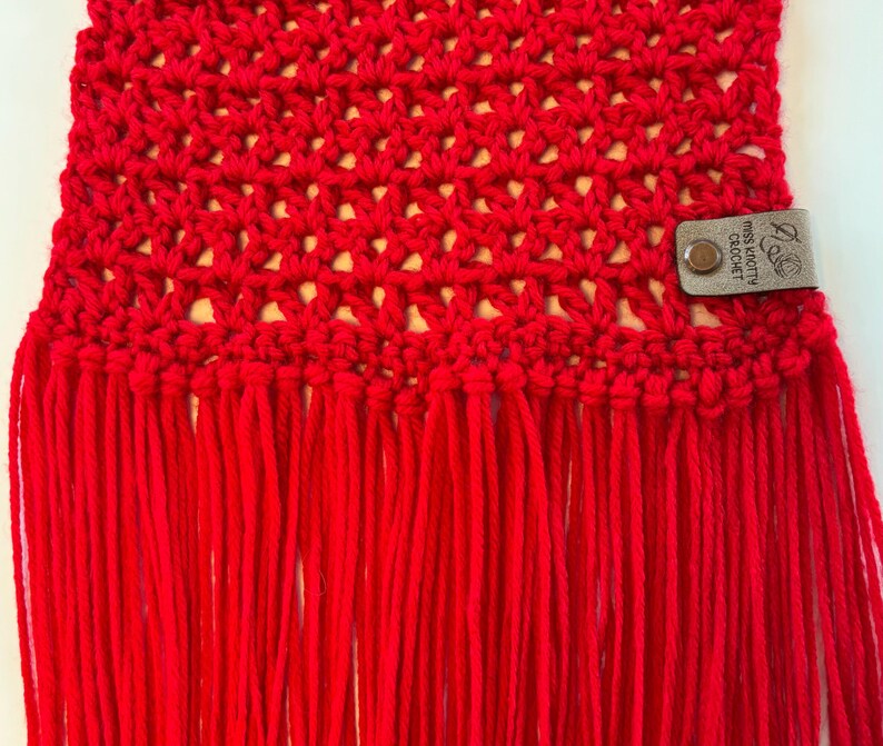 May include: A close-up of a vibrant red crocheted scarf with a fringe. The scarf features an open weave pattern. A small leather tag with the text "Miss Pretty Crochet" is attached to the scarf.