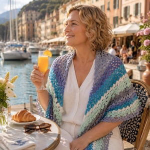 May include: A triangular crochet shawl featuring a gradient of colors including shades of blue, teal, and white. The shawl has an openwork, lacy stitch pattern with a scalloped edge, draped over a white garment. The design is lightweight and textured.
