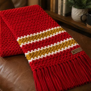 May include: A red crocheted scarf with white and gold stripes. The scarf has a fringed edge and a small leather tag. The scarf is folded on a brown leather surface.