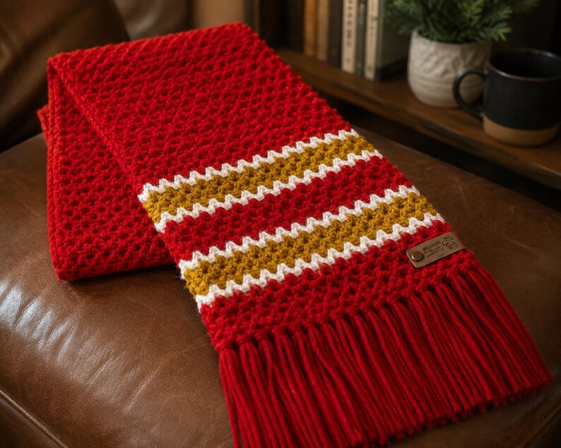 May include: A red crocheted scarf with white and gold stripes. The scarf has a fringed edge and a small leather tag. The scarf is folded on a brown leather surface.