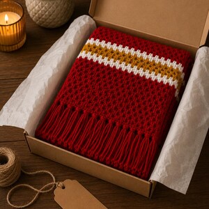 May include: A red crocheted scarf with white and gold stripes and fringe, presented in a cardboard box with tissue paper. A lit candle, a small plant, and a tag with twine are on a wooden surface.