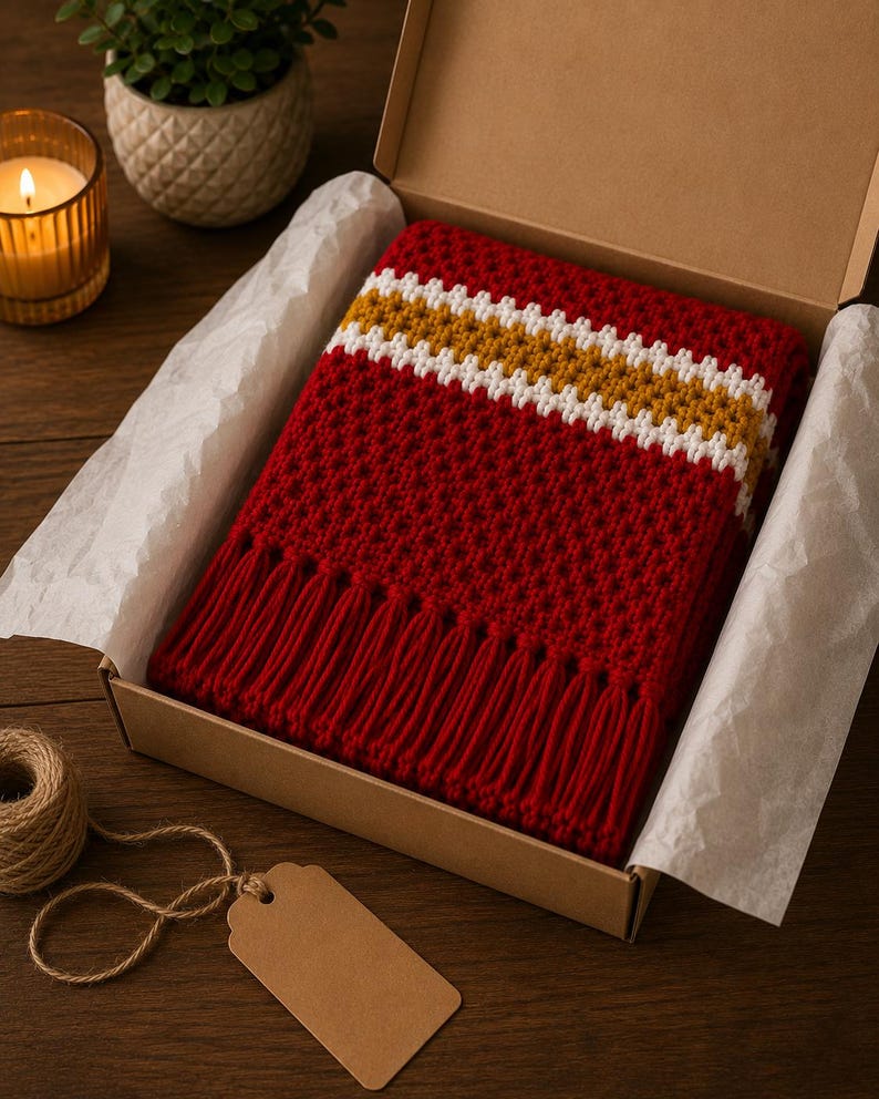 May include: A red crocheted scarf with white and gold stripes and fringe, presented in a cardboard box with tissue paper. A lit candle, a small plant, and a tag with twine are on a wooden surface.