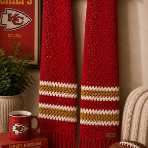 May include: A red crochet scarf with white and gold stripes and fringe, hanging on a wooden clothes hanger. The scarf is displayed with a mug and books featuring the Kansas City Chiefs logo. A football is also present.