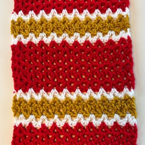 May include: A hand-crocheted scarf with a striking design of red, white, and gold stripes. The scarf's textured stitch highlights the detailed craftsmanship. The colour combination creates a bold and eye-catching accessory, perfect for adding a pop of colour to any outfit.