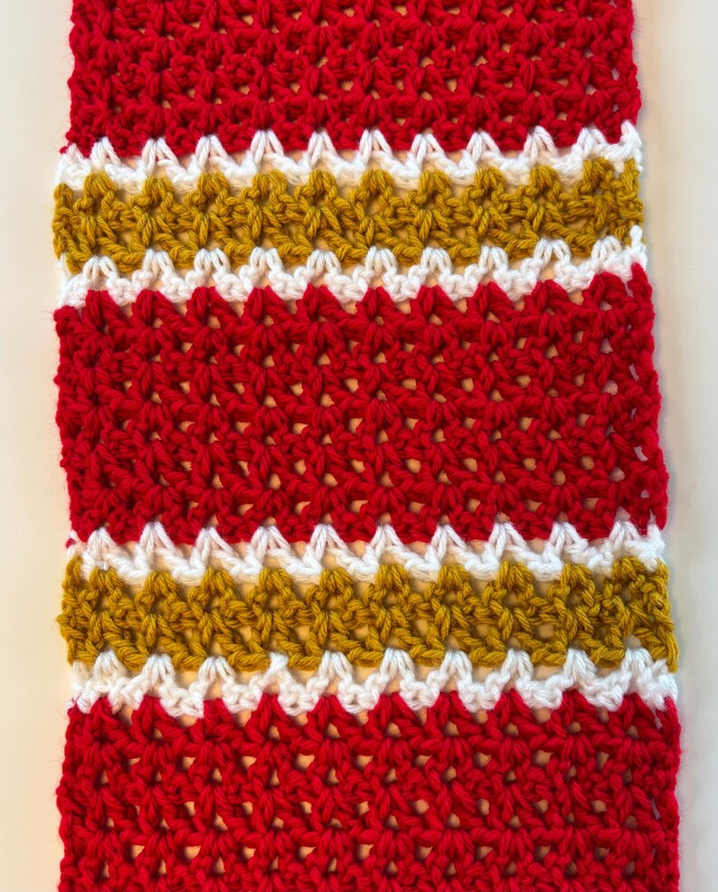 May include: A hand-crocheted scarf with a striking design of red, white, and gold stripes. The scarf's textured stitch highlights the detailed craftsmanship. The colour combination creates a bold and eye-catching accessory, perfect for adding a pop of colour to any outfit.