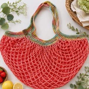 May include: A coral-colored crochet market bag with a wide, open-mesh body and two integrated handles. The upper trim and handles feature a variegated yarn pattern in shades of blue, yellow, and green. A small brown leather-like tag is attached.