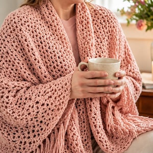 May include: A dusty pink crochet wrap featuring an openwork diamond pattern and long fringe trim along the edges. The garment is draped over the shoulders and arms, showcasing a textured, lightweight yarn construction with a soft, matte finish.