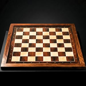 Puede incluir: Un tablero de ajedrez de madera hecho a mano con un marco marrón oscuro y una superficie de juego a cuadros con cuadrados claros y oscuros alternados. El tablero está sobre una superficie negra, mostrando la veta de la madera y la artesanía.