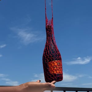 Könnte beinhalten: Ein roter, gehäkelter Weinflaschenhalter vor einem strahlend blauen Himmel. Der Halter umschließt eine dunkle Flasche mit einem orangefarbenen Etikett. Die Flasche wird von einer Hand gehalten.