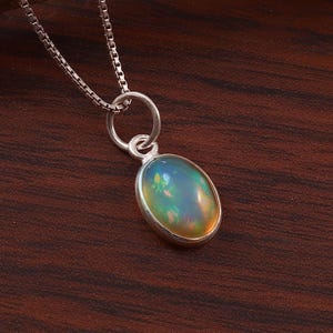 May include: A silver box chain necklace featuring a single oval-shaped cabochon opal pendant set in a smooth silver bezel. The opal displays iridescent flashes of green, orange, and yellow colors against a translucent, milky white base stone.