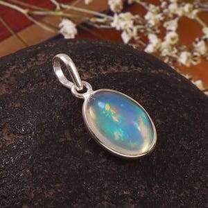 Puede incluir: Un colgante de forma ovalada con una piedra preciosa de ópalo iridiscente y lisa, engastada en un marco de metal pulido de tono plateado. La piedra muestra una base azul translúcida con destellos de luz verde, amarilla y naranja en su superficie.