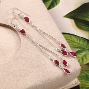 May include: A pair of long silver drop earrings featuring multiple marquise-cut red gemstones. Each earring consists of a thin silver wire hook connected to a vertical silver chain that suspends three distinct red marquise-shaped stones.