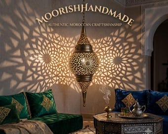 May include: A brass pendant lamp with intricate cut-out patterns hangs from the ceiling, casting geometric shadows on the wall. The text MOORISHHANDMADE AUTHENTIC MOROCCAN CRAFTSMANSHIP is visible above the lamp in the centre of the image.