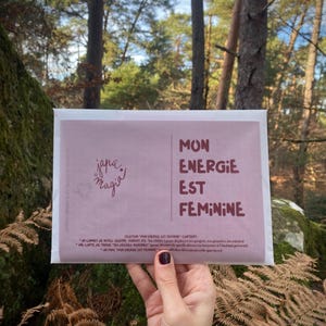 Peut inclure: Une enveloppe rose avec les mots "MON ENERGIE EST FEMININE" en texte bordeaux. L'enveloppe porte le logo "japa magia" dans le coin supérieur gauche. L'arrière-plan est une scène de forêt avec des arbres et des fougères.