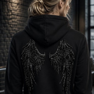 May include: A black hooded sweatshirt featuring a large, detailed graphic of angel wings on the back. The wings are rendered in a dark, textured style with numerous small, reflective embellishments that create a shimmering effect.