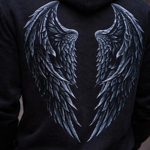 May include: Dark gray hoodie featuring a detailed, grayscale depiction of angel wings on the back. The wings are intricately designed with feathers, creating a striking visual contrast against the dark fabric. The hoodie has a hood and long sleeves.
