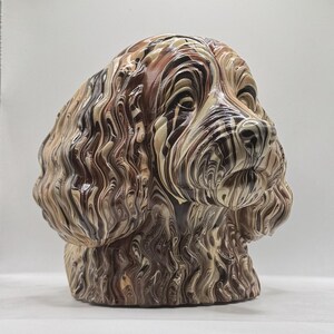 May include: A sculpted dog head figurine featuring a marbled pattern of swirling brown, cream, and black colours. The sculpture displays detailed textures representing long, wavy fur across the ears and neck, with a smooth, rounded nose and eyes.