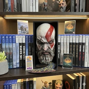 Puede incluir: Una estantería de madera oscura muestra carcasas de videojuegos y un busto de un personaje con una marca roja en la cabeza. Los juegos incluyen "God of War", "Elden Ring" y "Spider-Man". Un hacha decorativa está frente al busto.