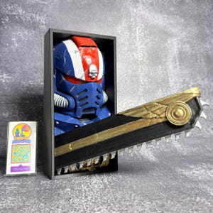 May include: A blue, red, and white helmet with a skull emblem is displayed in a black box. A black and gold chainsaw is positioned in front of the helmet. The helmet and chainsaw are likely part of a cosplay or collectible item.