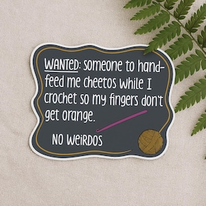May include: A dark gray sticker with a white border and gold trim. The text reads "WANTED: someone to hand-feed me cheetos while I crochet so my fingers don't get orange. NO WEIRDOS." A crochet hook and yarn ball are also shown.