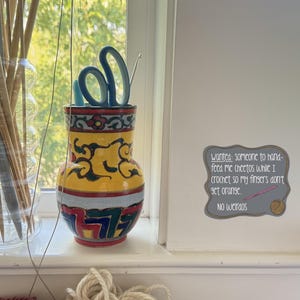 May include: A decorative vase with scissors and a crochet hook, next to a glass jar of knitting needles. The vase is yellow with a colorful design. A sign reads "Wanted: someone to hand-feed me cheetos while I crochet so my fingers don't get orange. No weirdos."
