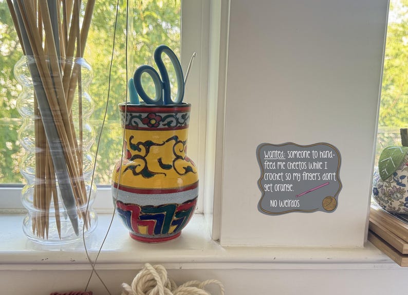 May include: A decorative vase with scissors and a crochet hook, next to a glass jar of knitting needles. The vase is yellow with a colorful design. A sign reads "Wanted: someone to hand-feed me cheetos while I crochet so my fingers don't get orange. No weirdos."