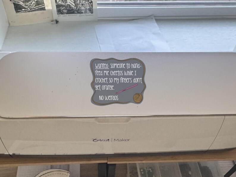 May include: A white Cricut Maker machine with a sticker that reads "Wanted: someone to hand-feed me Cheetos while I crochet so my fingers don't get orange. No weirdos." The sticker has a gray border and a pink crochet hook.