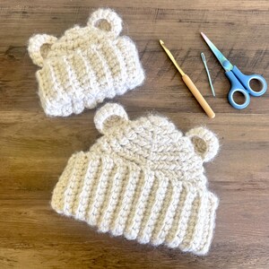 May include: Two handmade, light beige crochet bear hats with ear details. A crochet hook, needle, and blue scissors are also visible. The hats are made with a ribbed stitch and are likely for babies or children.