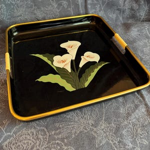 May include: A square black serving tray with a gold-colored rim and light-colored woven accents on the side handles. The center of the tray features a graphic illustration of three white calla lily flowers with green leaves on a black background.