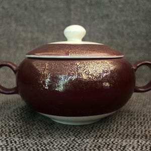 May include: A maroon ceramic sugar bowl with a lid and two handles. The bowl and lid are decorated with a gold floral pattern. The lid has a white knob. The bowl sits on a white base.