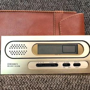 Peut inclure: Un réveil Seiko Melodic Alarm vintage avec un cadran doré et un étui en cuir marron. L'horloge comprend un affichage numérique, un haut-parleur et des boutons de commande. Le texte "SEIKO MELODIC ALARM" est imprimé sur le cadran.