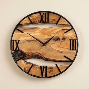 Puede incluir: Un reloj de pared redondo con una esfera de madera natural y números romanos negros. Las manecillas y el marco exterior son negros. La veta de la madera es visible, añadiendo un toque rústico al diseño. El reloj mide aproximadamente 30 cm de diámetro.