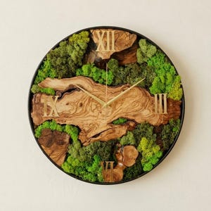 May include: Round wall clock with a black frame, featuring a natural wood and moss design. The clock face has gold Roman numerals and hands. The wood has a unique grain pattern, and the moss is a vibrant green.