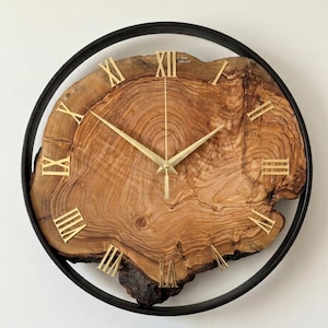 May include: A circular wall clock with a natural wood face, showcasing the tree's grain. Gold Roman numerals and hands are set against the wood, enclosed by a black metal frame. The clock's design emphasizes the wood's natural texture.