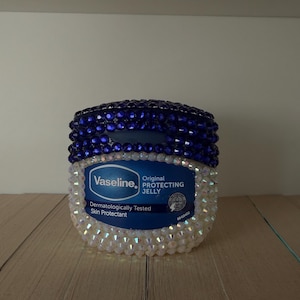 May include: A jar of Vaseline Original Protecting Jelly, decorated with blue and white crystals. The lid is adorned with blue crystals, while the base features white crystals. The jar is displayed on a wooden surface.