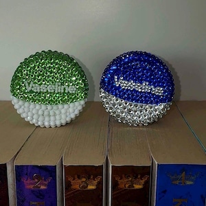 May include: Two decorative jar lids, one green and one blue, each encrusted with rhinestones. The green lid has white beads at the base, and the blue lid has silver beads. Both lids have the word "Vaseline" in white script.