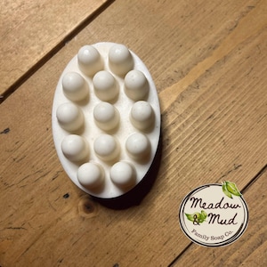 May include: Oval white soap with massage nodules. The soap is on a wooden surface. A circular logo with the text "Meadow & Mud Family Soap Co." is in the lower right corner.