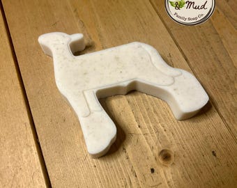 Puede incluir: Una pastilla de jabón blanca con forma de cordero y textura moteada. El jabón está sobre una superficie de madera, y un logotipo circular con el texto "Meadow & Mud Family Soap Co." es visible en la esquina superior derecha. El jabón es de color claro y neutro.