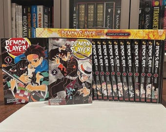 Puede incluir: Una colección de volúmenes de manga "Demon Slayer", incluyendo un set de caja completo. Las portadas presentan ilustraciones coloridas de personajes de la serie. Las lomos son negros con texto blanco. Los libros están dispuestos sobre una superficie blanca.