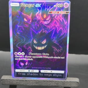 May include: A holographic Gengar ex Pok&eacute;mon trading card. The card features a purple and black illustration of Gengar with the text "Phantom Gate" and "from shadow to mega abyss". The card has a HP of 220 and is an Ultra Beast.